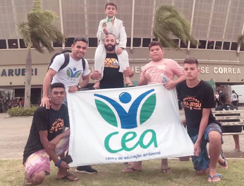 Foto de pessoas segurando uma bandeira do CEA em frente a um estádio. Um menino vestindo kimono está segurando uma medalha.