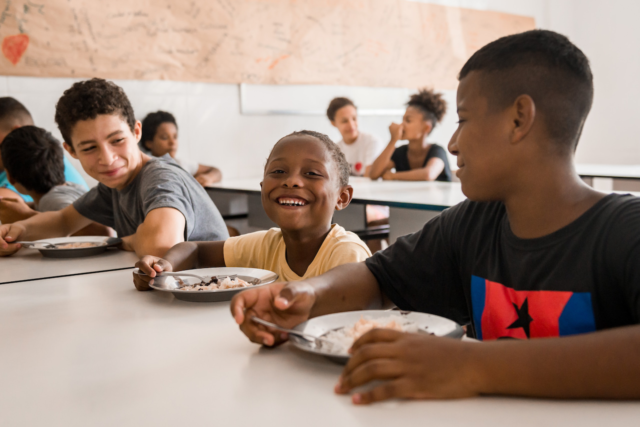 Meninos comendo sentados à mesa. O menino do meio sorri para a câmera