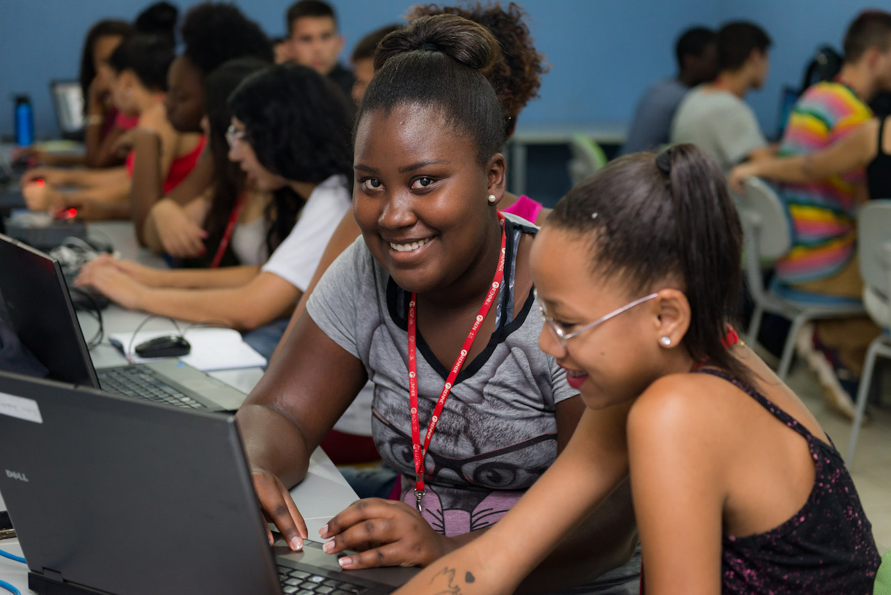 Sala com diversos jovens trabalhando em computadores. A imagem foca em duas jovens